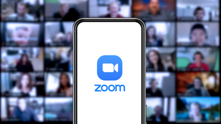 Zoom-sovelluksen ikoni matkapuhelimessa, ja taustalla kokoukseen osallistuvien henkilöiden kuvat.