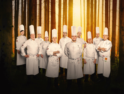 Suomen kokkimaajoukkue seisoo kokkiasut päällä kellertävän taustan edessä.